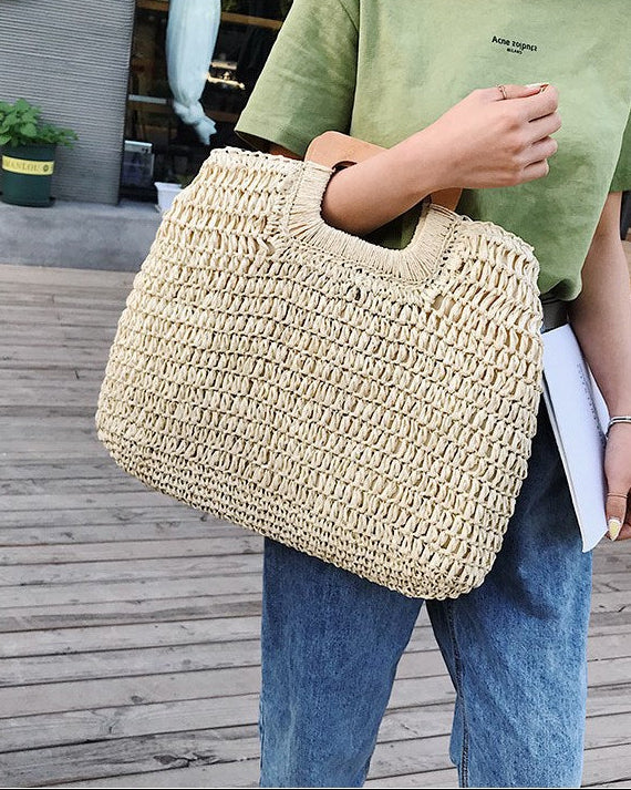 Person holding a woven beige bag outdoors on a wooden deck.