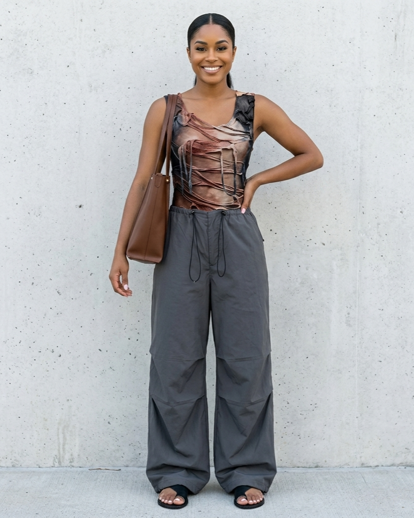 Woman wearing a sheer top and gray pants against a white wall