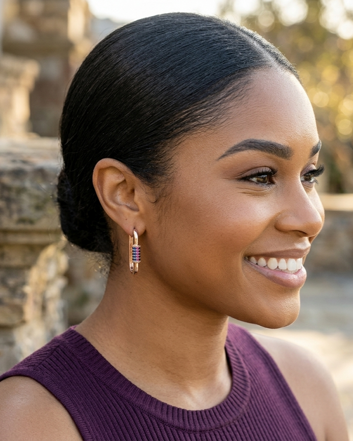 Woman with styled hair and earrings, wearing a purple top, outdoors.