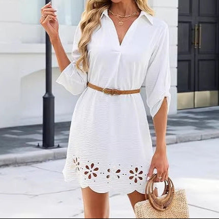 Woman in a white dress with floral cutouts, straw hat, and sunglasses standing in front of a building.