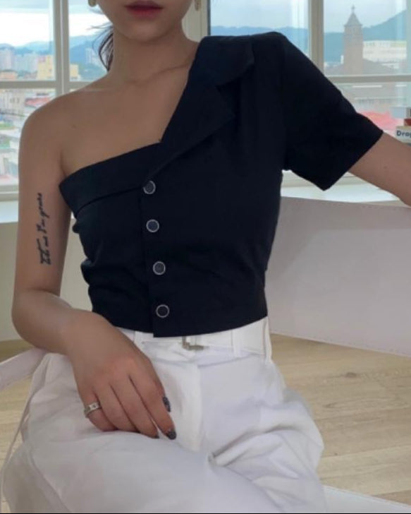 Woman wearing a black one-shoulder top and white pants sitting in a room with large windows.