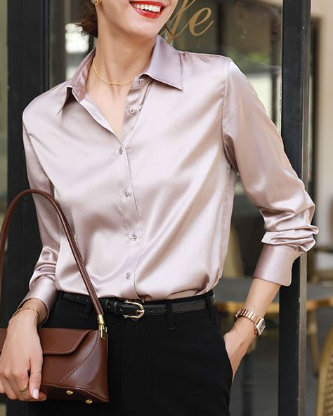 Woman in a light pink blouse and black pants holding a brown handbag, standing in front of a store window.