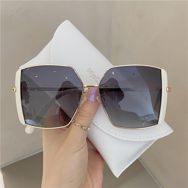 Sunglasses with gold frame and dark lenses held by a hand, with a white case in the background.