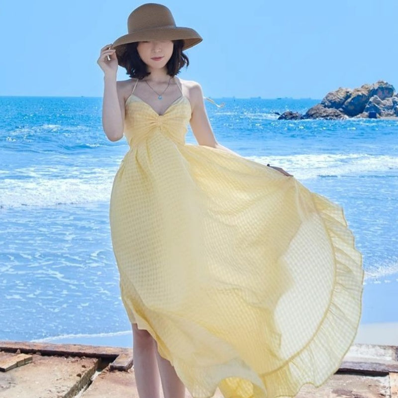 Woman in a yellow dress and brown hat standing on a beach with blue water and rocks in the background.