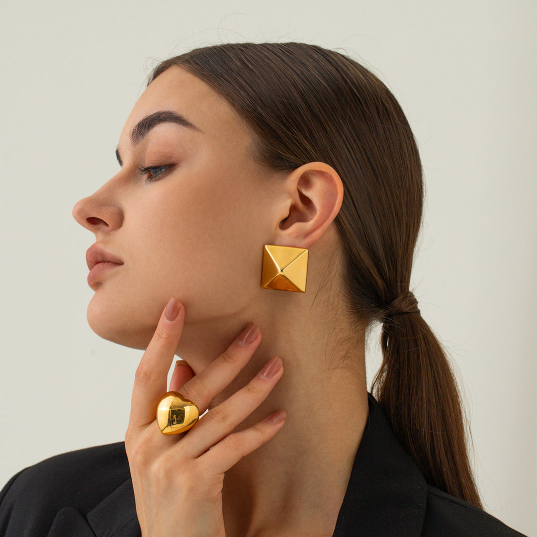 Woman wearing gold pyramid-shaped earrings and a matching ring on a neutral background
