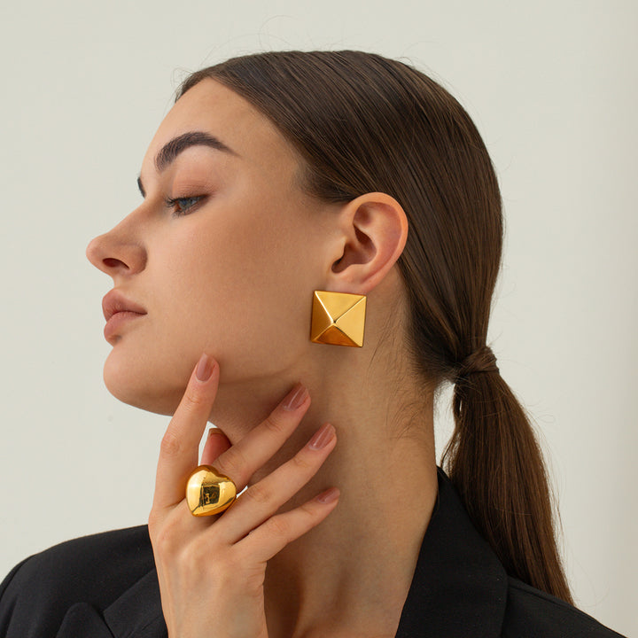 Woman wearing gold pyramid-shaped earrings and a matching ring on a neutral background